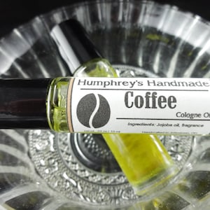 May include: Two small glass bottles of coffee-scented cologne oil. The bottles have black roll-on tops and labels that say "Humphrey's Handmade Coffee Cologne Oil". The label also includes a coffee bean graphic and the ingredients: "Jojoba oil, fragrance".