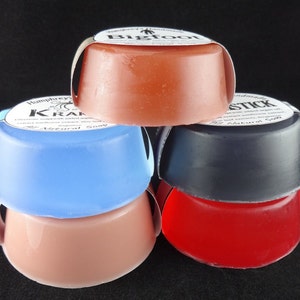 May include: Four bars of handmade soap stacked on top of each other. The top bar is a light orange color with the text "Bigfoot" on the label. The second bar is light blue with the text "Humphrey's Krazy" on the label. The third bar is dark gray with the text "Stick" on the label. The bottom bar is red.