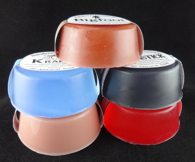 May include: Four bars of handmade soap stacked on top of each other. The top bar is a light orange color with the text "Bigfoot" on the label. The second bar is light blue with the text "Humphrey's Krazy" on the label. The third bar is dark gray with the text "Stick" on the label. The bottom bar is red.