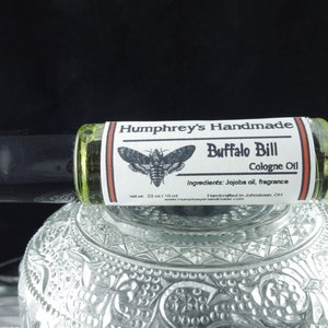 May include: A glass roll-on bottle with a white label that says "Humphrey's Handmade Buffalo Bill Cologne Oil" with a black and white moth illustration. The label also lists the ingredients as "Jojoba oil, fragrance" and says "Handcrafted in Johnstown, OH".