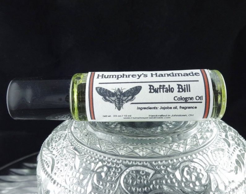 May include: A glass roll-on bottle with a white label that says "Humphrey's Handmade Buffalo Bill Cologne Oil" with a black and white moth illustration. The label also lists the ingredients as "Jojoba oil, fragrance" and says "Handcrafted in Johnstown, OH".
