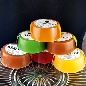 Pu&ograve; includere: Sei saponette rotonde fatte a mano impilate su un piatto di vetro trasparente. I saponi sono di vari colori, tra cui arancione, verde, marrone, rosso e giallo. Ogni saponetta ha un'etichetta con il nome del profumo e le parole "Sapone artigianale".