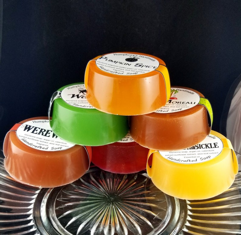 Pu&ograve; includere: Sei saponette rotonde fatte a mano impilate su un piatto di vetro trasparente. I saponi sono di vari colori, tra cui arancione, verde, marrone, rosso e giallo. Ogni saponetta ha un'etichetta con il nome del profumo e le parole "Sapone artigianale".