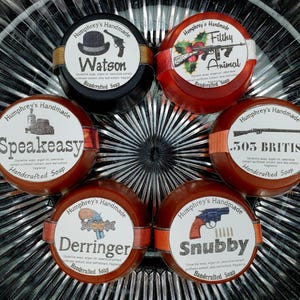 May include: A collection of handcrafted soaps with various labels. The labels feature illustrations of a hat and gun, a machine gun with holly, a building, a gun, a pistol, and a revolver. The soaps are round and have a brown or black base.