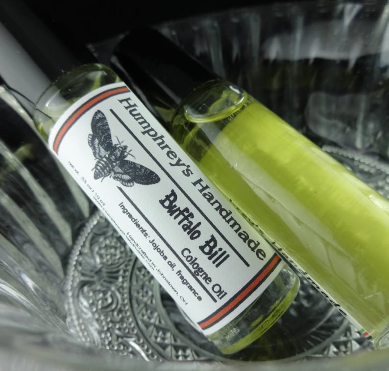 May include: Two small glass bottles of cologne oil with black caps. The label on one bottle reads "Humphrey's Handmade Buffalo Bill Cologne Oil" with a moth graphic. The ingredients are listed as "Jojoba oil, fragrance".