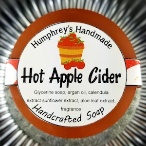 May include: A round, orange soap bar with a white label that reads "Humphrey's Handmade Hot Apple Cider Glycerine soap, argan oil, calendula extract sunflower extract, aloe leaf extract, fragrance Handcrafted Soap". The label features a red and white illustration of a basket of apples.