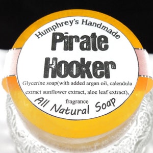 Pu&ograve; includere: Un sapone rotondo giallo con un'etichetta bianca che recita "Humphrey's Handmade Pirate Hooker Glycerine soap(with added argan oil, calendula extract sunflower extract, aloe leaf extract), fragrance All Natural Soap"