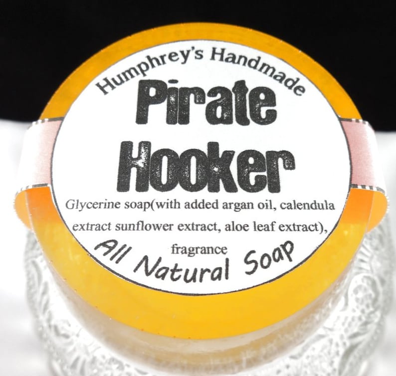 Pu&ograve; includere: Un sapone rotondo giallo con un'etichetta bianca che recita "Humphrey's Handmade Pirate Hooker Glycerine soap(with added argan oil, calendula extract sunflower extract, aloe leaf extract), fragrance All Natural Soap"
