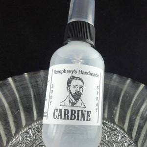May include: A clear glass bottle with a black spray top and a white label. The label says "Humphrey's Handmade Body Spray" and "CARBINE". A black and white illustration of a man with a beard is on the label.