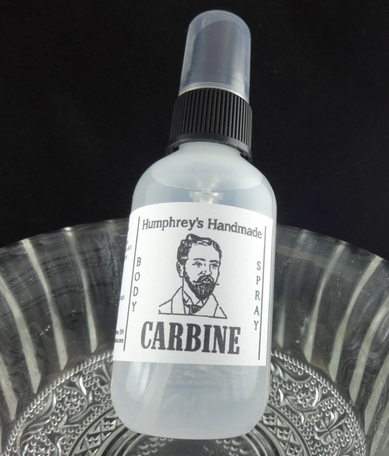 May include: A clear glass bottle with a black spray top and a white label. The label says "Humphrey's Handmade Body Spray" and "CARBINE". A black and white illustration of a man with a beard is on the label.