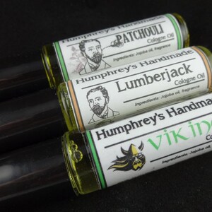 May include: Three glass roll-on bottles with black caps. Each bottle has a green, white and black label with a different design and text. The labels read "Humphrey's Handmade Patchouli Cologne Oil", "Humphrey's Handmade Lumberjack Cologne Oil", and "Humphrey's Handmade Viking Cologne Oil".