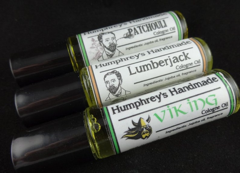 May include: Three glass roll-on bottles with black caps. Each bottle has a green, white and black label with a different design and text. The labels read "Humphrey's Handmade Patchouli Cologne Oil", "Humphrey's Handmade Lumberjack Cologne Oil", and "Humphrey's Handmade Viking Cologne Oil".