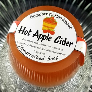May include: A round, orange-colored soap bar with a white label that reads "Humphrey's Handmade Hot Apple Cider Handcrafted Soap". The label features a basket of red apples and the text "Glycerine soap, argan oil, calendula extract sunflower extract, aloe leaf extract, fragrance".