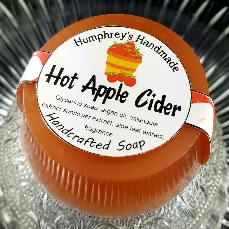 May include: A round, orange-colored soap bar with a white label that reads "Humphrey's Handmade Hot Apple Cider Handcrafted Soap". The label features a basket of red apples and the text "Glycerine soap, argan oil, calendula extract sunflower extract, aloe leaf extract, fragrance".