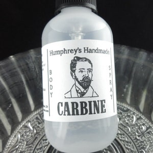 May include: A clear glass bottle with a black spray top and a white label. The label features a black and white illustration of a man with a beard and the text "Humphrey's Handmade Body Spray CARBINE".
