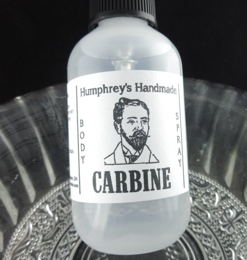 May include: A clear glass bottle with a black spray top and a white label. The label features a black and white illustration of a man with a beard and the text "Humphrey's Handmade Body Spray CARBINE".