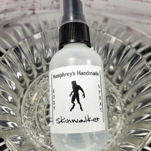 May include: A clear body spray bottle with a black pump top. The label reads "Humphrey's Handmade BODY SPRAY skinwalker" with a black silhouette of a creature. The bottle is in a clear glass bowl.