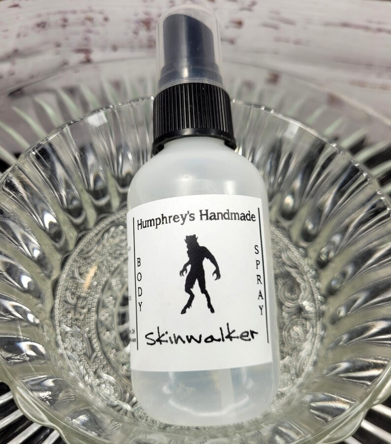 May include: A clear body spray bottle with a black pump top. The label reads "Humphrey's Handmade BODY SPRAY skinwalker" with a black silhouette of a creature. The bottle is in a clear glass bowl.