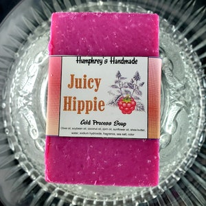 May include: A pink bar of soap with a label that reads "Humphrey's Handmade Juicy Hippie Cold Process Soap". The label also lists the ingredients: olive oil, soybean oil, coconut oil, corn oil, sunflower oil, shea butter, water, sodium hydroxide, fragrance, sea salt, and color.