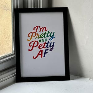 May include: A framed print with the text "I'm Pretty and Petty AF" in a rainbow gradient font. The print is in a black frame and is displayed on a white surface.