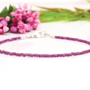 Könnte beinhalten: Ein zartes, magentafarbenes Armband mit Perlen und einem silbernen Verschluss. Das Armband besteht aus kleinen, quadratisch geschliffenen Perlen. Rosa Blüten und grünes Laub befinden sich im Hintergrund.