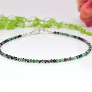 May include: A delicate bracelet made of small, round beads in green, black, and pink. The bracelet is strung on a thin, silver-coloured chain and has a simple clasp. The jewellery is displayed on a white surface.