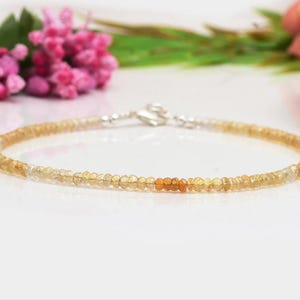 Peut inclure: Un bracelet délicat composé de petites perles rondes jaune doré. Les perles changent de couleur, avec quelques perles orange plus foncées au centre. Le bracelet a un fermoir argenté et est présenté sur une surface blanche.