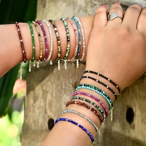 Puede incluir: Una colección de pulseras de cuentas en varios colores, incluyendo rojo, verde, morado, blanco, marrón, turquesa y azul. Las pulseras están ensartadas en delicadas cadenas y se muestran en muñecas.