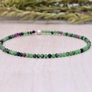May include: A delicate bracelet composed of small, round beads in shades of green, black, and pink. The beads are strung together, forming a circular shape. The bracelet is displayed on a light-colored surface, with greenery in the background.