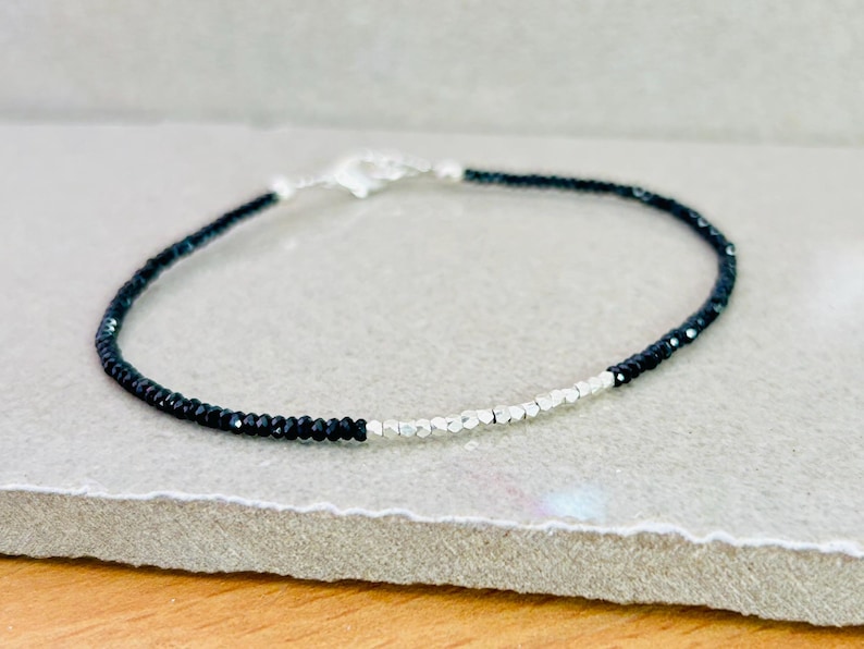 May include: A delicate bracelet featuring small, black beads and silver-colored beads. The bracelet is strung on a thin, flexible cord and has a silver-colored clasp. The jewelry is displayed on a light gray surface.