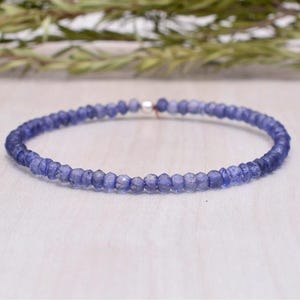 May include: A delicate bracelet composed of small, faceted, blue-violet gemstones. The beads are strung together with a silver-colored clasp. The bracelet is displayed on a light-colored surface, with a blurred green background.