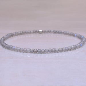 Puede incluir: Una delicada pulsera compuesta por pequeñas cuentas redondas grises. Las cuentas tienen sutiles destellos azules e iridiscentes. La pulsera está ensartada en un cordón fino y flexible y asegurada con un pequeño cierre plateado. Joyería.
