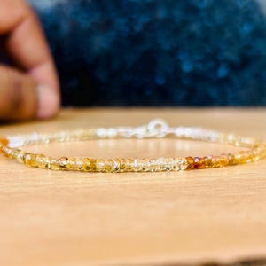 May include: Delicate beaded bracelet featuring small, round, yellow and orange gemstones. The bracelet has a silver clasp and is displayed on a light brown surface. The gemstones transition in color from light yellow to a deeper orange.