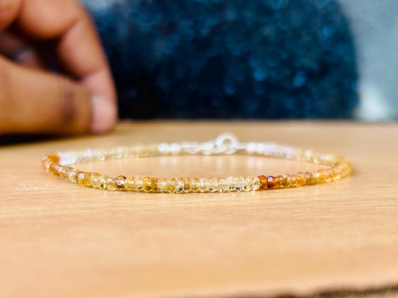 May include: Delicate beaded bracelet featuring small, round, yellow and orange gemstones. The bracelet has a silver clasp and is displayed on a light brown surface. The gemstones transition in color from light yellow to a deeper orange.