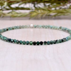 May include: A delicate bracelet composed of small, faceted emerald beads. The beads are a mix of dark and light green, strung together on an elastic band. A small silver bead is visible. The bracelet is displayed on a light wooden surface.