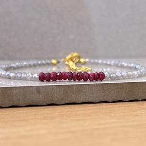 Peut inclure: Un bracelet délicat composé d'un fil de petites perles claires et facettées, avec une rangée centrale de perles de rubis rouge foncé. Le bracelet a un fermoir doré. Le bijou est présenté sur une surface aux tons neutres.