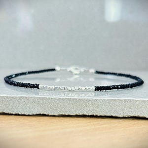 May include: A delicate bracelet featuring alternating sections of black and silver beads. The bracelet is secured with a silver clasp. The beads are small and evenly spaced, creating a minimalist and elegant design. The bracelet is displayed on a neutral surface.