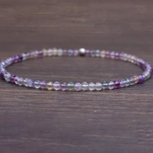 May include: A delicate bracelet composed of small, faceted beads in shades of purple, green, and clear. The beads are strung together, forming a flexible band. The bracelet is displayed on a dark wooden surface.