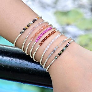 Puede incluir: Una colección de pulseras de cuentas en varios colores, incluyendo rosa, marrón y verde, apiladas en una muñeca. Cada pulsera presenta pequeñas cuentas coloridas ensartadas en una banda plateada. Las pulseras se muestran sobre un fondo borroso de vegetación.