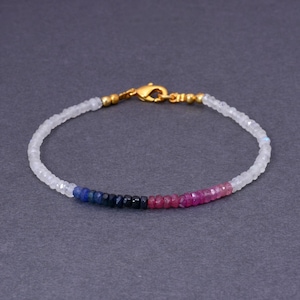 Pode incluir: Uma pulseira delicada com uma corrente de pequenas contas facetadas em um gradiente de cores. A pulseira transita do branco para o azul, preto e depois para tons de rosa e vermelho. Um fecho dourado prende a pulseira.