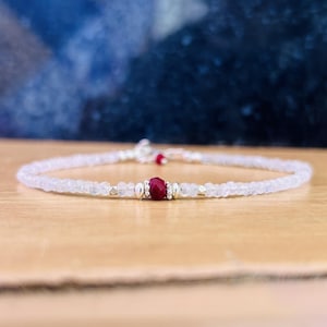 May include: Delicate bracelet featuring small, round, white beads and silver accents. A vibrant, faceted, red gemstone is the focal point, complemented by smaller red beads. The bracelet is displayed on a wooden surface.