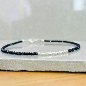 May include: A delicate bracelet featuring alternating sections of small, dark beads and silver-colored beads. The bracelet is secured with a silver clasp. The jewelry is displayed on a light gray surface.