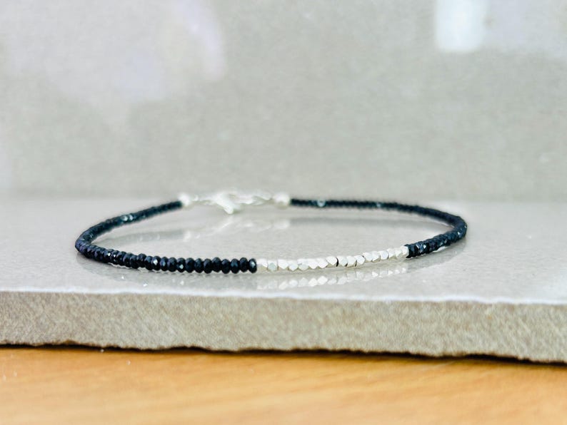 May include: A delicate bracelet featuring alternating sections of small, dark beads and silver-colored beads. The bracelet is secured with a silver clasp. The jewelry is displayed on a light gray surface.