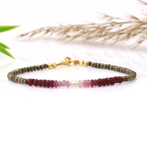 May include: A delicate bracelet featuring a mix of gemstones. The bracelet has a gold clasp and is strung with small, faceted beads in shades of dark green, red, pink, and clear. The gemstones are arranged in a gradient pattern.