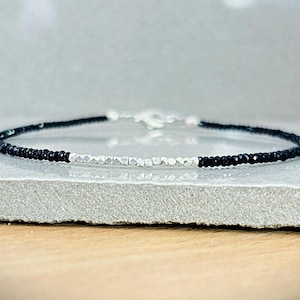 May include: A delicate bracelet featuring small, black beads and silver-toned, faceted beads. The bracelet has a simple design with a silver clasp. The jewelry is displayed on a light gray surface.
