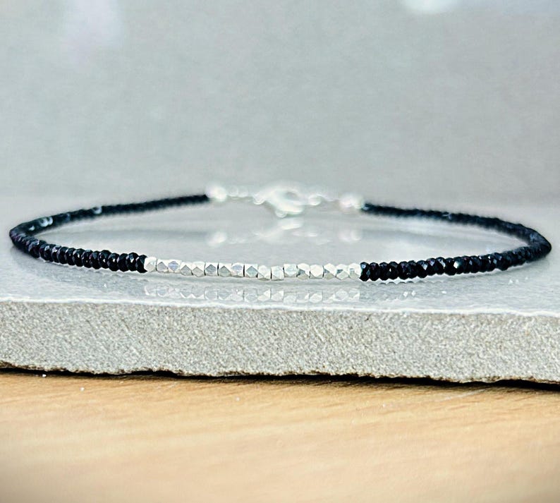 May include: A delicate bracelet featuring small, black beads and silver-toned, faceted beads. The bracelet has a simple design with a silver clasp. The jewelry is displayed on a light gray surface.