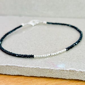 May include: A delicate bracelet featuring small, faceted black beads and silver-colored beads. The bracelet is strung on a thin, flexible cord and has a silver-colored clasp. The jewelry is displayed on a neutral-toned surface.