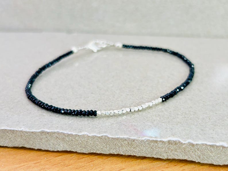 May include: A delicate bracelet featuring small, faceted black beads and silver-colored beads. The bracelet is strung on a thin, flexible cord and has a silver-colored clasp. The jewelry is displayed on a neutral-toned surface.