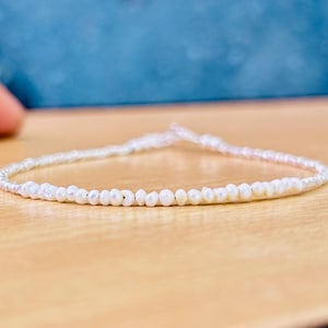Könnte beinhalten: Zartes Armband aus kleinen, weißen Perlen. Die Perlen sind auf einem dünnen, weißen Band aufgefädelt. Die Perlen haben unterschiedliche Größen und Formen. Das Armband wird auf einer hellbraunen Oberfläche präsentiert. Die Perlen haben einen Durchmesser von ca. 0,3 cm.
