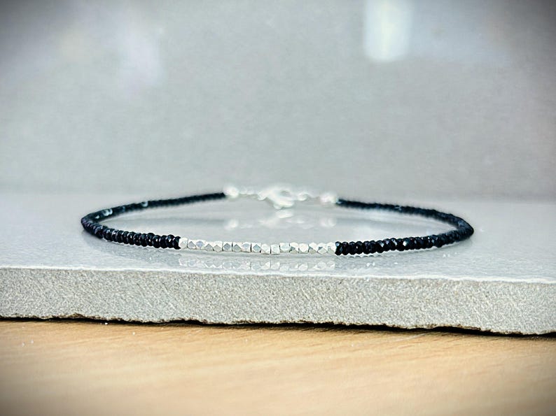 May include: A delicate bracelet featuring alternating sections of black and silver beads. The bracelet has a silver clasp and is displayed on a neutral surface. The beads are small and evenly spaced, creating a minimalist design.